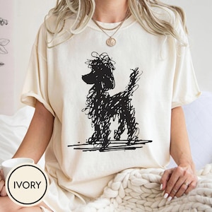 May include: Ivory-coloured t-shirt with a black scribble-style poodle illustration. The shirt has short sleeves and a relaxed fit. The word "IVORY" is printed on a white circle in the lower left corner. The shirt is made of a soft, comfortable material.