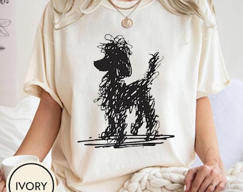 May include: Ivory-colored t-shirt featuring a black scribble-style poodle illustration. The shirt has short sleeves and a relaxed fit. The word "IVORY" is printed on a white circle in the lower left corner. The shirt is made of a soft, comfortable material.