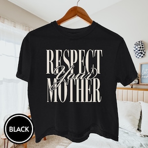 May include: A black short-sleeve t-shirt featuring the text "RESPECT YOUR MOTHER" printed in a large, cream-colored serif font across the chest. The word "Your" is written in a cursive script style, while "Respect" and "Mother" are in block letters.