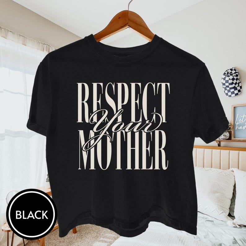 May include: A black short-sleeve t-shirt featuring the text "RESPECT YOUR MOTHER" printed in a large, cream-colored serif font across the chest. The word "Your" is written in a cursive script style, while "Respect" and "Mother" are in block letters.