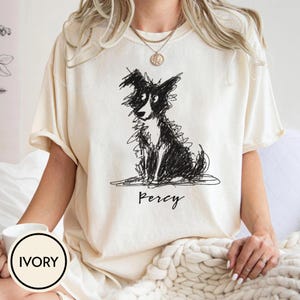 May include: An ivory short-sleeved t-shirt featuring a black ink-style sketch of a dog sitting down. Below the dog illustration, the name Percy is printed in a black cursive script. The shirt has a crew neckline and a relaxed fit.