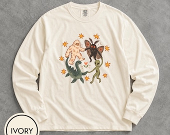May include: An ivory long-sleeve t-shirt featuring a graphic of a Bigfoot, a mothman, a Loch Ness monster, and an alien holding hands in a circle around a small flower, surrounded by orange stars. The shirt has a Comfort Colors brand label.