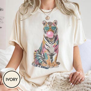May include: Ivory-colored t-shirt featuring a colorful tiger wearing sunglasses and blowing a bubblegum bubble. The tiger has a rainbow-colored design. The word "IVORY" is printed on a white circle.