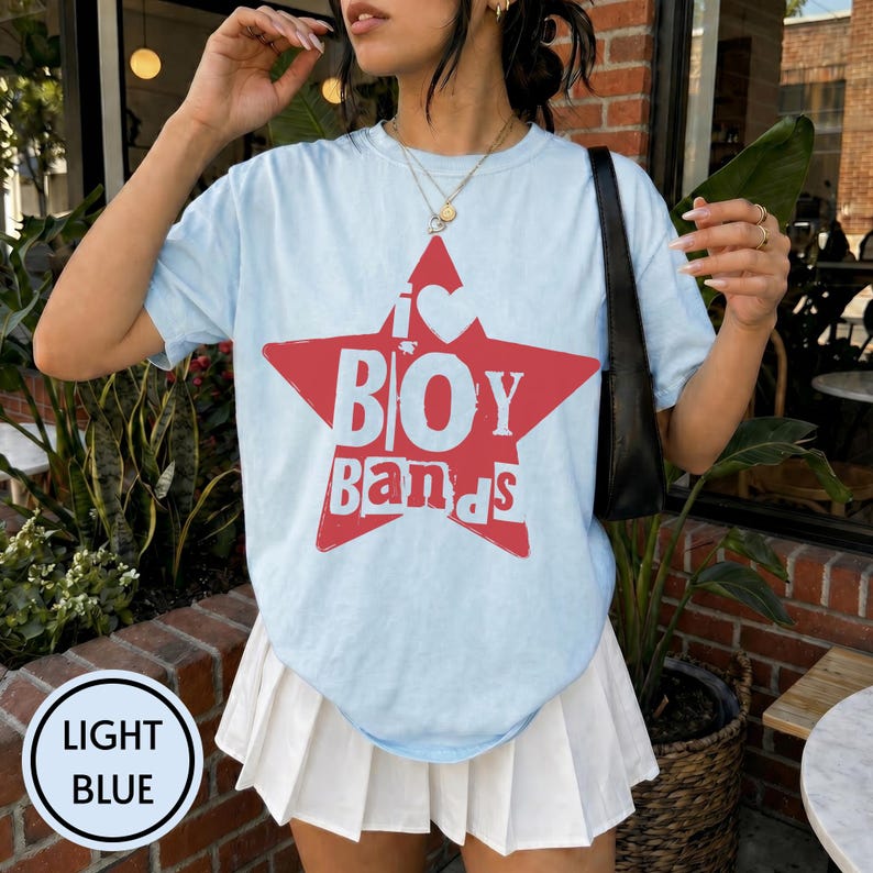 Puede incluir: Una camiseta azul claro con un gr&aacute;fico de estrella roja grande y el texto blanco I LOVE BOY BANDS, combinada con una falda plisada blanca y un bolso de hombro negro. La camiseta tiene cuello redondo, mangas cortas y un collar dorado en capas.