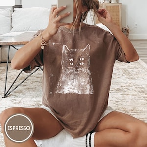 May include: A brown short-sleeved t-shirt featuring a white distressed graphic print of a cat with four eyes. The shirt has a crew neckline and a loose, relaxed fit. A circular label in the bottom corner displays the text ESPRESSO.