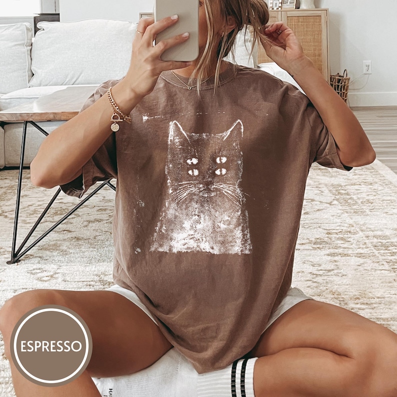 May include: A brown short-sleeved t-shirt featuring a white distressed graphic print of a cat with four eyes. The shirt has a crew neckline and a loose, relaxed fit. A circular label in the bottom corner displays the text ESPRESSO.
