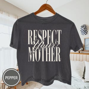 May include: A charcoal gray short-sleeve t-shirt featuring the white text "RESPECT YOUR MOTHER" printed on the front. The word "RESPECT" and "MOTHER" are in a tall, bold serif font, while "Your" is in a cursive script font.