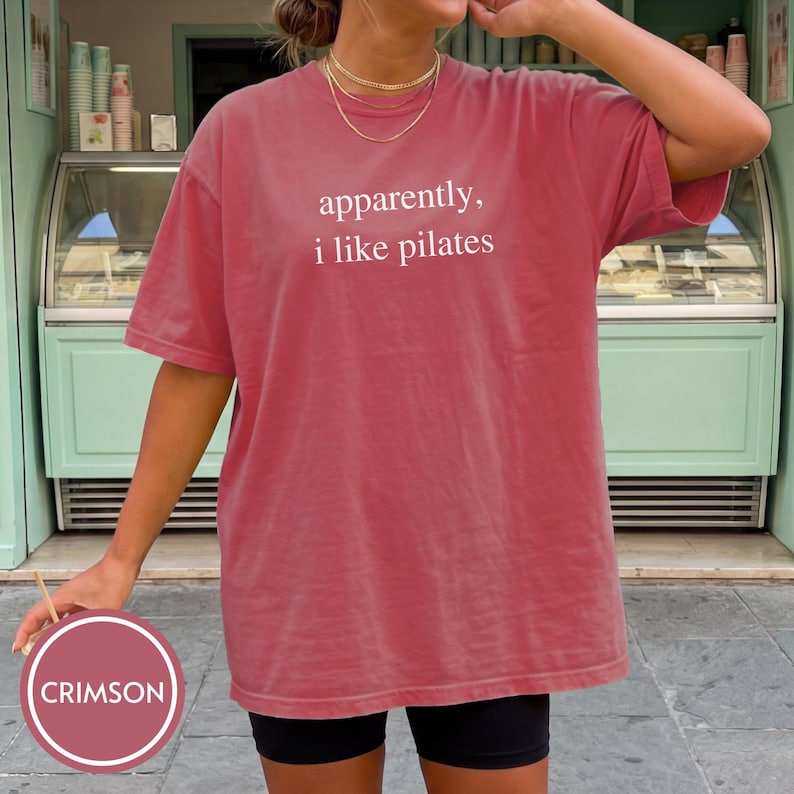 May include: Crimson-colored t-shirt with the text "apparently, i like pilates" in white. The shirt is a short-sleeved, crew neck style. The model is wearing black shorts. The background includes a mint green storefront.