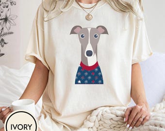 May include: Ivory-colored t-shirt featuring a graphic of a greyhound dog with a blue sweater adorned with white stars and red trim. The shirt has short sleeves and a round neckline. The word "IVORY" is printed on a white circle.