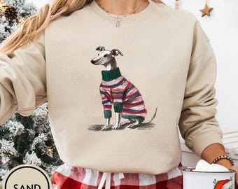 May include: A beige sweatshirt featuring a detailed illustration of a greyhound dog wearing a striped sweater in red, green, and blue. The dog is sitting, with its tail curled. The word "SAND" is printed on a circular label.