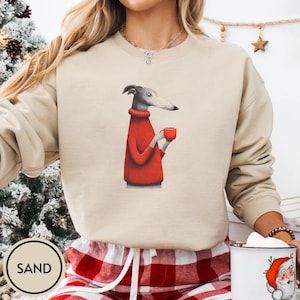 May include: Sand-colored sweatshirt featuring a greyhound dog wearing a red sweater and holding a red mug. The dog is depicted in a whimsical, illustrated style. The word "SAND" is printed on a circular label at the bottom.