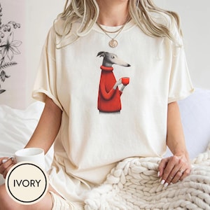 May include: Ivory-colored t-shirt featuring a greyhound dog wearing a red sweater and holding a red mug. The dog illustration is centered on the shirt. The shirt has short sleeves and a relaxed fit. A white mug with the word "IVORY" is in the foreground.