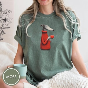 May include: Green t-shirt featuring a cartoon greyhound dog wearing a red sweater and holding a red cup. The shirt has rolled-up sleeves and a relaxed fit. The word "MOSS" is in a green circle in the lower left corner.