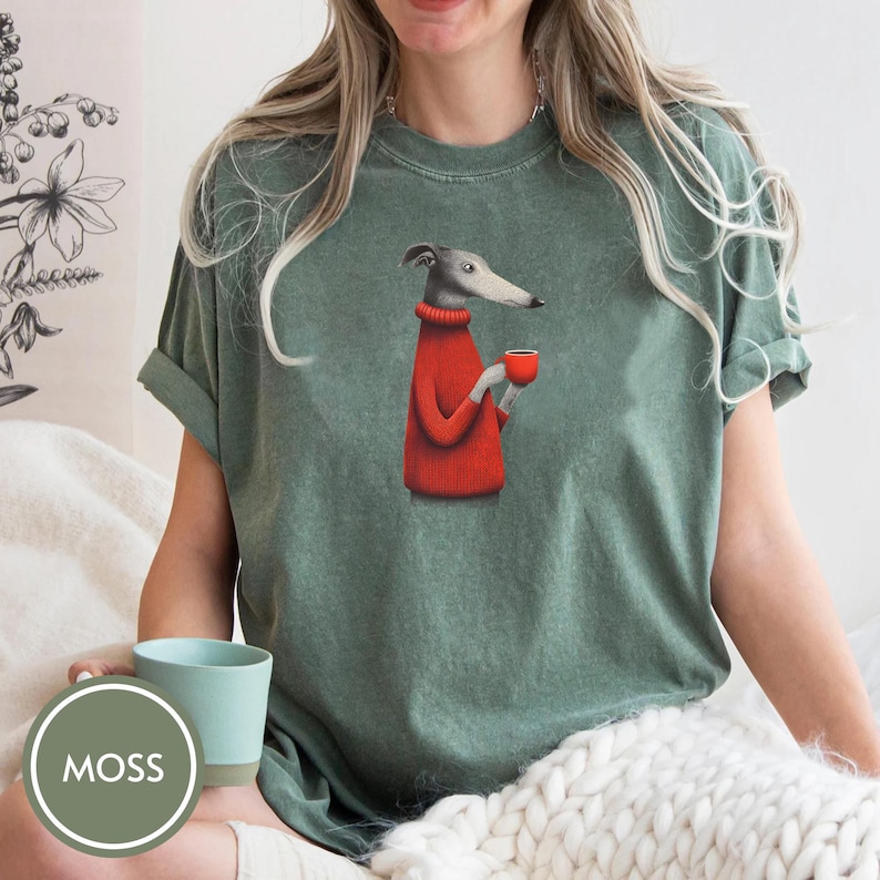 May include: Green t-shirt featuring a cartoon greyhound dog wearing a red sweater and holding a red cup. The shirt has rolled-up sleeves and a relaxed fit. The word "MOSS" is in a green circle in the lower left corner.