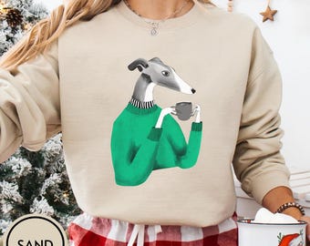 May include: Sand-colored sweatshirt featuring a greyhound dog illustration. The dog is depicted wearing a green sweater and holding a small grey mug. The sweatshirt has a crew neck and long sleeves. The word "SAND" is printed on a circular label.