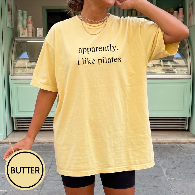 May include: A yellow t-shirt with the text "apparently, i like pilates" in black. The shirt is a light yellow color and has a relaxed fit. The word "BUTTER" is in a circle in the bottom left corner.