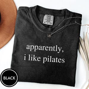 May include: Black t-shirt with the text "apparently, i like pilates" in white. The shirt has a crew neck and a small tag on the upper back. A silver necklace with a pendant is draped on the shirt.