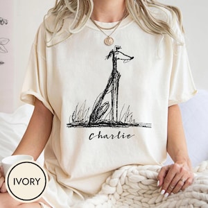 Può includere: Una t-shirt a maniche corte color avorio con un'illustrazione nera in stile schizzo di un cane alto e snello seduto nell'erba. Sotto il cane, il nome Charlie è stampato in un carattere corsivo nero. La maglietta ha un girocollo.
