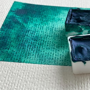 May include: Close-up of watercolor paints and a painted swatch. Two white square paint containers are filled with dark blue paint. A teal and green watercolor wash is on a textured white paper.