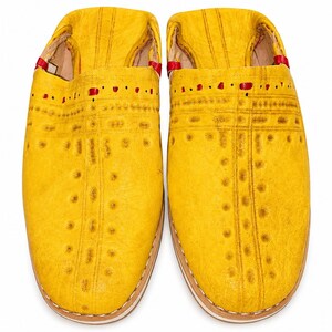 Könnte beinhalten: Ein Paar gelbe Lederschuhe mit dekorativen Nähten und kleinen Punktmustern. Die Schuhe haben eine runde Spitze und eine kontrastierende weiße Sohle. Rote Akzente sind innen und um die Öffnung sichtbar.