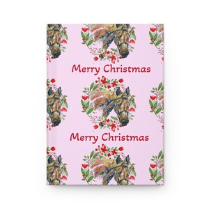 May include: A pink journal with a repeating pattern of watercolor horse heads, wreaths, and the text "Merry Christmas" in red. The horse heads are brown and tan, and the wreaths feature red flowers and green leaves.