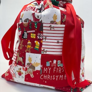 May include: A festive Christmas gift bag in red and white, with a drawstring closure and red ribbon ties. The patchwork design includes reindeer, penguins, and the text "MY FIRST CHRISTMAS".