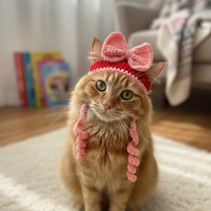 Pode incluir: Um gato ruivo usando um chapéu vermelho de crochê com um laço rosa e tranças rosa combinando. O gato está sentado em um tapete branco. O texto "Pawsome Knits - Handmade with Love!" está na parte inferior da imagem.