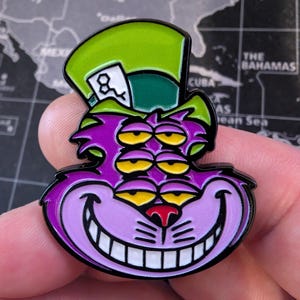 May include: A purple and green enamel pin featuring a stylized Cheshire Cat design with multiple yellow eyes and a wide grin. The cat wears a green top hat with a white card. The pin has a black outline.