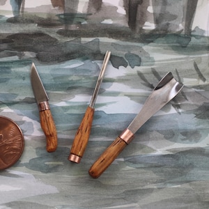 May include: Three small wood-handled carving tools with copper accents are displayed next to a penny. The tools have stainless steel blades of different shapes. The handles are made of light-colored wood.