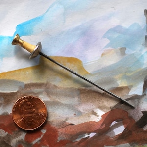 May include: A miniature metal fencing foil with a gold-colored handle and a silver-colored guard, positioned diagonally next to a copper-colored United States one-cent coin for scale, resting against a multi-colored abstract watercolor background.