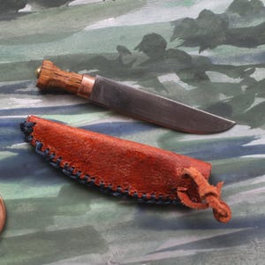 May include: A small, handcrafted knife with a wooden handle and a metal blade, resting beside a brown leather sheath with blue stitching. A penny is next to the knife for size comparison.