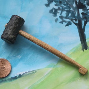May include: A miniature hammer with a dark, textured head and a light wooden handle. A penny is next to the hammer for scale. The background features a watercolor painting of a landscape with a tree and rolling hills.