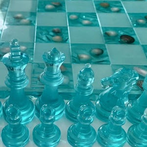 Può includere: Un set di scacchi turchese con pezzi trasparenti. La scacchiera presenta un motivo a scacchi blu e bianco con conchiglie. I pezzi degli scacchi includono re, regine, alfieri, cavalli, torri e pedoni.