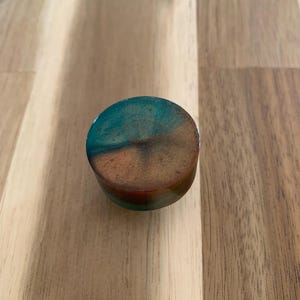 May include: A small, round object with a flat top and sides. The top is a blend of teal and brown hues, with a speckled texture. The object appears to be made of resin or a similar material, and it sits on a wooden surface.