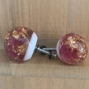 Peut inclure: Deux boutons de meuble décoratifs. Chaque bouton est une sphère transparente avec une teinte rouge-rosé, incrustée de flocons de feuilles d'or. Une bande blanche entoure la base de chaque bouton. Les boutons sont fixés à des vis argentées.