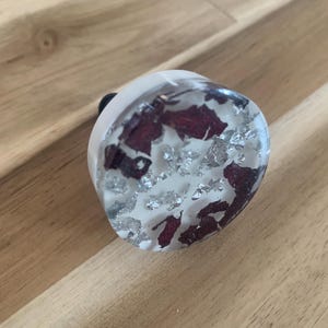 May include: A clear, rounded knob with embedded red and silver accents. The knob has a white base and a black stem. The decorative element is encased in clear resin, creating a unique and artistic design. This knob is suitable for furniture or cabinet doors.