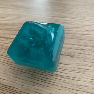 May include: A teal resin cube with a shimmering interior, resting on a light brown wooden surface. The translucent square has rounded edges and internal bubbles. The object's colour is a vibrant teal, and it appears to be handmade.