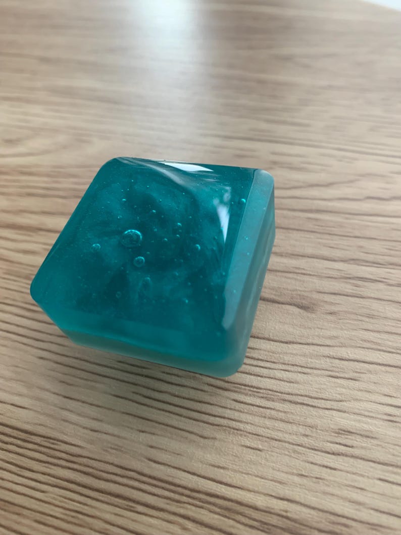 May include: A teal resin cube with a shimmering interior, resting on a light brown wooden surface. The translucent square has rounded edges and internal bubbles. The object's colour is a vibrant teal, and it appears to be handmade.