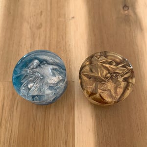 May include: Two circular decorative items. One is blue with silver swirls, the other is gold with a crumpled foil effect. Both are set against a wooden surface. The objects appear to be resin or glass.