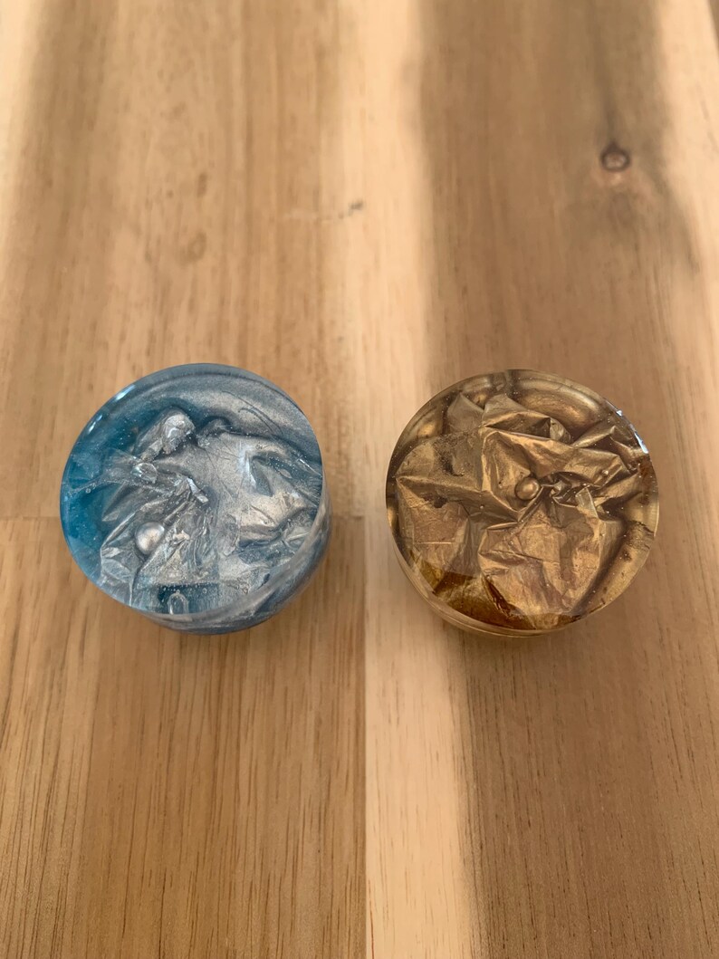 May include: Two circular decorative items. One is blue with silver swirls, the other is gold with a crumpled foil effect. Both are set against a wooden surface. The objects appear to be resin or glass.