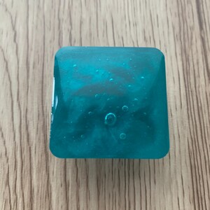 May include: A square, teal resin object with a marbled effect and bubbles. It sits on a wooden surface. The translucent resin allows light to pass through. The object's colour is a vibrant teal.