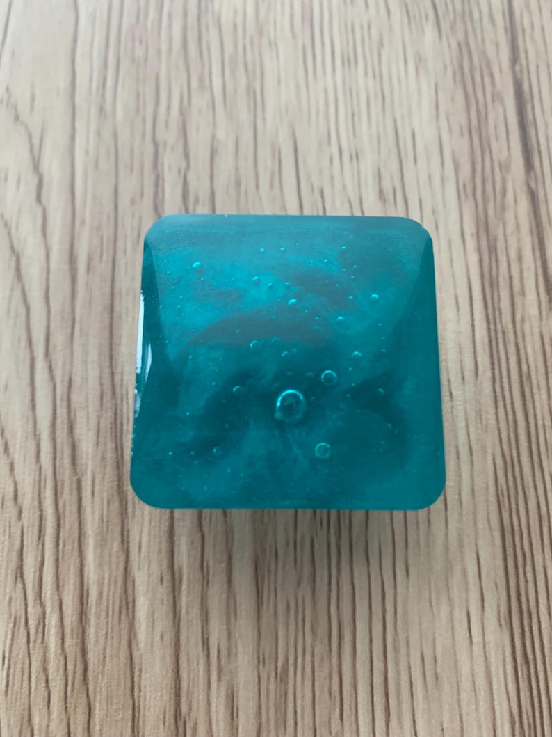 May include: A square, teal resin object with a marbled effect and bubbles. It sits on a wooden surface. The translucent resin allows light to pass through. The object's colour is a vibrant teal.