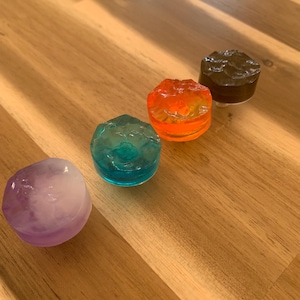 Op de afbeelding: Vier kleurrijke, ronde decoratieve objecten in tinten paars, blauwgroen, oranje en bruin. De objecten hebben een textuur, edelsteenachtig uiterlijk en zijn diagonaal op een houten oppervlak geplaatst. De objecten lijken van hars of glas te zijn.