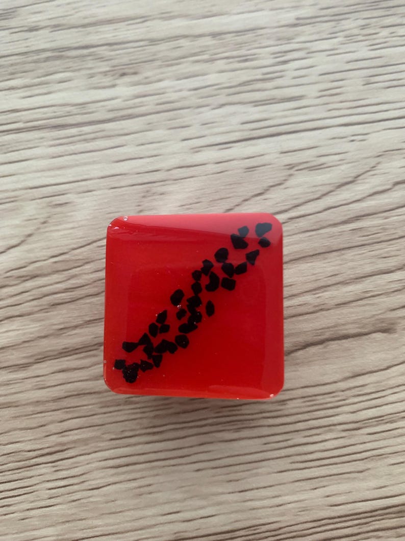 Pode incluir: Um objeto quadrado de resina vermelha com um padr&atilde;o diagonal de pequenas formas pretas. O objeto est&aacute; sobre uma superf&iacute;cie de madeira castanha clara.