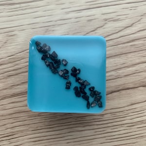 Pode incluir: Uma base para copos quadrada de resina azul clara com uma dispers&atilde;o de pequenas pedras cinza escuras. A base para copos est&aacute; sobre um fundo de madeira, mostrando sua superf&iacute;cie lisa e elementos decorativos embutidos. O design &eacute; simples e moderno.