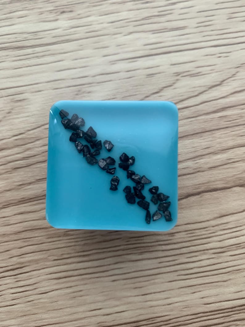 Pode incluir: Uma base para copos quadrada de resina azul clara com uma dispers&atilde;o de pequenas pedras cinza escuras. A base para copos est&aacute; sobre um fundo de madeira, mostrando sua superf&iacute;cie lisa e elementos decorativos embutidos. O design &eacute; simples e moderno.