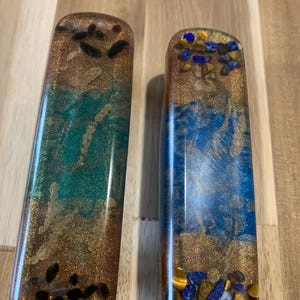 May include: Two rectangular resin objects with embedded stones and glitter. One features teal and gold tones with black stones, the other has blue and gold with blue, yellow, and gray stones. Both have a glossy finish.