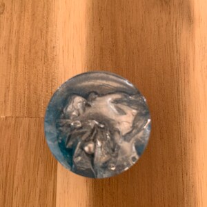 May include: A circular resin object in shades of blue and silver. The object features a translucent blue exterior and a swirling silver pattern within. It rests on a wooden surface.