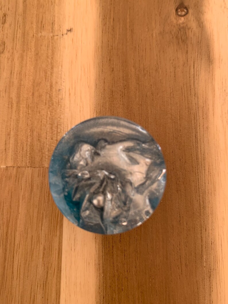 May include: A circular resin object in shades of blue and silver. The object features a translucent blue exterior and a swirling silver pattern within. It rests on a wooden surface.