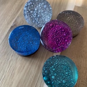 Op de afbeelding: Vijf kleurrijke, ronde oordopjes met een textuur, bubbelachtig ontwerp. De oordopjes zijn in tinten blauw, teal, paars, grijs en bruin. Ze worden getoond op een lichtbruin houten oppervlak.